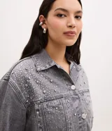 Campera de jean gris con efecto desteñido, corte oversized, cuello inglés, cierre frontal con botones niquelados y aplicaciones de brillos y piedras en la parte superior delantera.