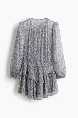 Vestido corto de tejido transparente, con estampado de patrón geométrico y floral en tonos azules y blancos, cuello redondo con pequeña abertura y tiras para atar. Tiene manga larga abullonada, cintura elastizada y falda con volados en capas.