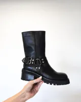 Bota de cuero negra con caña media, suela track y detalle de arnés con hebillas y tachas plateadas.