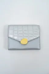Billetera pequeña de color plateado con solapa frontal texturizada efecto reptil y cierre decorativo con aplique circular amarillo.