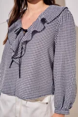 Blusa de corte oversize con estampado de cuadros vichy en blanco y negro. Presenta un cuello con volados, mangas largas con puños elásticos y un cierre frontal con lazo negro.