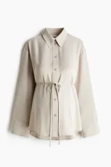 Camisa de sarga color beige, de corte holgado, con cuello clásico, botonadura frontal y mangas largas con puños anchos. Incorpora una tira fina para atar bajo el busto y bajo redondeado.