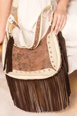 Cartera hobo de cuero color arena con apliques de gamuza chocolate y fantasía metalizada. Tiene flecos largos, tachas metálicas y herrajes plateados. Incluye correa corta fija y correa larga extensible y desmontable.