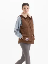 Chaleco matelaseado color marrón camel con canesú en corderoy color chocolate. Presenta cuello con corderito, cierre frontal con botones a presión, bolsillos con tapa y un parche bordado en el bolsillo derecho.