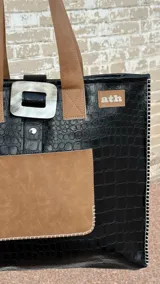 Tote bag de ecocuero negro con textura croco, cuenta con un bolsillo frontal de gamuza marrón y un detalle decorativo circular de guampa en el cierre superior. Posee doble asa de hombro y costuras visibles en los bordes.
