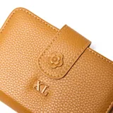 Billetera de tamaño mediano en color camel, confeccionada en material símil cuero con textura granulada. Presenta un cierre de solapa con broche decorado con una flor en relieve y el logo de la marca XL en la parte frontal.