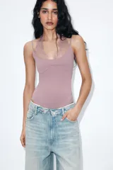 Body de tejido elástico color rosado con escote redondo y tirantes finos. Presenta un detalle de encaje a tono en la parte superior del escote que se ajusta mediante una lazada al cuello. Cuenta con cierre en el bajo mediante botones a presión.
