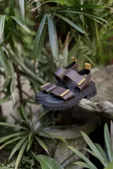 Sandalias de cuero marrón con suela track negra de plataforma. Presentan tiras ajustables con cierre de velcro y detalles en color mostaza en los extremos.
