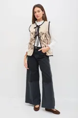 Chaleco de lana color beige con bordados florales artesanales en tonos marrones y blancos. Presenta un diseño sin mangas con volados en los hombros y cierre frontal mediante lazos negros ajustables.