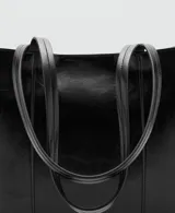 Bolso tipo shopper color negro, confeccionado en poliuretano con acabado brillante y efecto arrugado, con asas largas y costuras verticales decorativas en el frente.