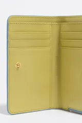 Billetera mediana con exterior rígido y textura brillante, con estampado de leopardo en tonos azul celeste, negro y naranja. Presenta cierre de cremallera y logo metálico de Bimba y Lola en el frente.