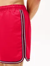 Short de baño rojo con cintura elástica ajustable con cordón blanco. Tiene dos bolsillos laterales y un bolsillo trasero de parche. El bajo presenta una franja con los colores azul marino, blanco y rojo.