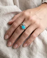 Anillo de plata 925 con diseño multicapa, decorado con una piedra central de ópalo sintético en tonos azules y verdes, y una piedra redonda translúcida más pequeña en la parte superior.