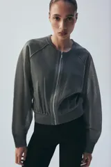 Chaqueta estilo bomber de punto color beige con acabado brillante, cuello redondo, manga larga con puños elásticos y cierre frontal con cremallera.