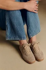 Zapatilla deportiva de gamuza color visón, con cordones a tono y suela de goma marrón. Presenta un diseño retro de running con el nombre de la marca grabado en dorado en el lateral.