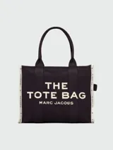 Bolso tote grande de tela negra con asas de mano y hombro. Presenta el texto "THE TOTE BAG MARC JACOBS" en color blanco en el frente. Los bordes laterales tienen una cinta blanca con texto repetido.