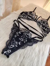 Conjunto de lencería compuesto por un bralette triangular y una tanga, confeccionados en encaje negro con detalles florales en color hueso. El bralette presenta breteles ajustables y diseño con tiras decorativas, mientras que la tanga cuenta con doble tira lateral regulable.