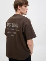 Remera marrón de manga corta con cuello redondo y diseño holgado, con un pequeño estampado tipográfico en el pecho.