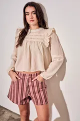 Blusa de color arena con diseño romántico, presenta volados en los hombros, detalles de encaje en el pecho y mangas largas con puños elásticos.