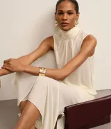 Blusa color crema sin mangas de crepé con cuello alto y lazo por tiras largas de tejido. La prenda tiene cierre por gota con detalle de perla artificial atrás del cuello y frunzidos en la parte delantera.