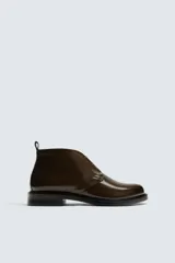 Bota de caña baja tipo chukka, confeccionada en piel con acabado brillante. Presenta cierre de cordones, horma redonda y suela a contraste con ligero tacón.