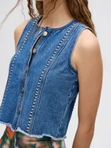 Chaleco de denim liviano con cierre frontal de botones metálicos, detalle de apliques metálicos verticales y ruedo con terminación desflecada.