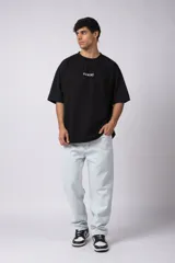 Remera negra de corte oversize confeccionada en tela pesada. Presenta un bordado frontal con la palabra 'REWIND' y un diseño de parche en la espalda con estética de papel roto.