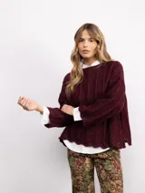Sweater de punto oversize en color bordó con destellos de lurex. Presenta un diseño acanalado vertical, cuello redondo y mangas largas con terminaciones acampanadas y bordes ondulados.