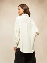 Camisa de pana color crema, con cuello camisero, botones negros en el frente y mangas largas abullonadas con puños.