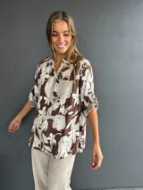 Blusa de corte holgado con estampado floral en tonos marrón y blanco. Presenta cuello camisero, cierre frontal con botones y un bolsillo en el pecho con detalle de lentejuelas. Las mangas son tres cuartos con trabas para ajustar.