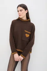 Sweater de punto color marrón con cuello alto, manga larga y calce relajado. Presenta detalles bordados en color mostaza en el cuello, puños, ruedo y bolsillo frontal, además de un aplique en forma de corazón en el bolsillo.