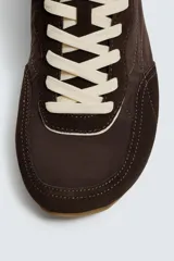 Zapatilla deportiva estilo barefoot, color marrón, con diseño de puntera ancha y redondeada. Confeccionada en una combinación de materiales textiles y detalles en serraje. Cuenta con suela fina de goma flexible y cordones en color crema.