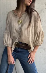 Camisa oversize color beige, con cuello redondo fruncido, cierre frontal de botones y mangas abullonadas con puños elásticos.