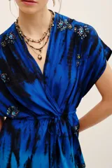 Vestido corto azul con estampado tie-dye en tonos negros y azules, escote en V cruzado, mangas cortas y cordón ajustable en la cintura. Presenta detalles de aplicaciones bordadas con lentejuelas en hombros y falda.