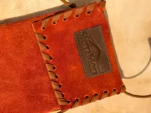 Funda para celular de gamuza azul marino con correa de cuero marrón y aplique de cuero marrón con logo de la marca.