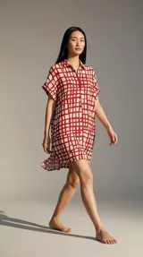 Vestido camisero de corte holgado y silueta relajada, con cuello camisero, cierre frontal de botones y mangas cortas. Presenta un estampado geométrico de cuadrícula en color rojo sobre fondo blanco.