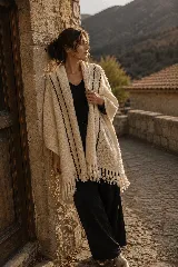 Ruana tejida a mano en lana merino, de color crudo con detalles de líneas verticales en color oscuro a contraste. Presenta un diseño envolvente con terminaciones de flecos en el ruedo.