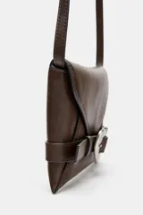 Bolso formato bandolera de material sintético con acabado metálico plateado. Presenta un diseño de solapa frontal con un cinturón decorativo que se cierra con una hebilla metálica en forma de corazón. Incluye un asa bandolera fina de cadena y un bolsillo interior con cierre.