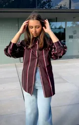 Camisa de corte oversize y manga larga, con diseño de rayas verticales en tonos bordó, blanco y negro. Presenta cuello clásico y cierre frontal con botones.