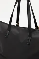 Cartera tipo tote de nylon negro, con diseño amplio y liviano. Presenta asas superiores fijas con detalles de tiras regulables y herrajes metálicos dorados. Incluye cierre principal y un pequeño logo metálico en la parte inferior frontal.