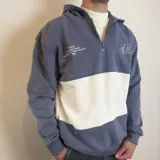 Canguro con capucha y cierre frontal parcial, de diseño color block en azul grisáceo y blanco. Presenta bordado con logo y texto en el pecho.