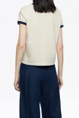 Remera corta de corte ajustado, color beige claro con finas rayas horizontales oscuras. Posee cuello redondo y ribetes en contraste de color azul marino en el cuello y los bordes de las mangas cortas. Lleva un pequeño bordado en forma de corazón con texto en el pecho.