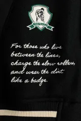 Campera estilo varsity con cuerpo negro y mangas en contraste color crema. Presenta múltiples parches y bordados decorativos en el frente y las mangas, cuello, puños y ruedo acanalados a rayas, y cierre frontal con botones a presión.