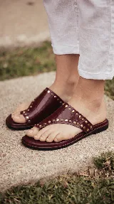 Sandalias chatas de cuero color bordó, con diseño de tira ancha que envuelve el empeine y sujeta el dedo gordo, decoradas con tachas metálicas doradas.