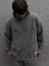Buzo oversize de cuello redondo en color gris, con diseño estampado en el frente que incluye logotipos y texto. Presenta puños y ruedo acanalados, con una pequeña etiqueta decorativa en la parte inferior.