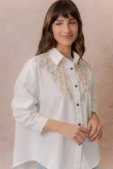 Camisa de denim azul con bordados florales en tonos amarillos y celestes sobre los hombros y el pecho. Presenta cuello clásico, cierre frontal con botones y mangas largas con puños.