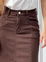 Pollera larga de tiro alto confeccionada en denim color marrón, con diseño ajustado en la parte superior y corte acampanado hacia el ruedo. Presenta costuras verticales decorativas y un acabado desflecado en el borde inferior.