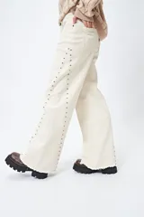 Pantalón de denim corte wide leg color marrón, con ruedo desflecado y detalle de tachas metálicas a lo largo de las costuras laterales.