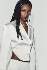 Blazer crop satinada de color blanco, con solapas, manga larga, cierre frontal con botones y detalle de abertura en la espalda.