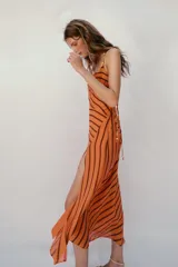Vestido midi de color naranja con rayas diagonales marrones, escote fluido y tirantes finos cruzados en la espalda con abalorios. Presenta corte evasé con abertura frontal y forro interior.